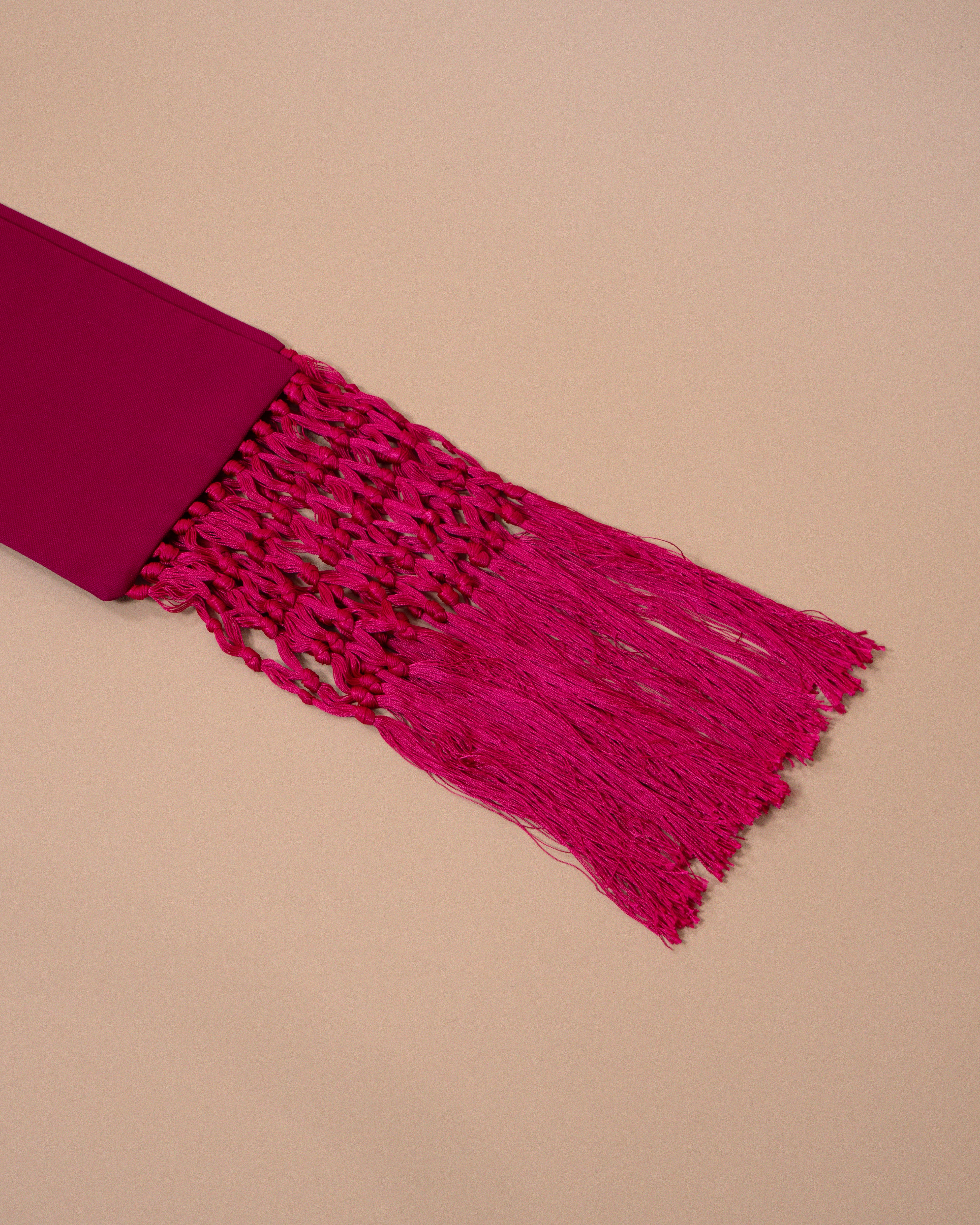 A close-up of a bright magenta pink woven textile accessory with a solid fabric end and long fringe, laid diagonally on a warm beige background.