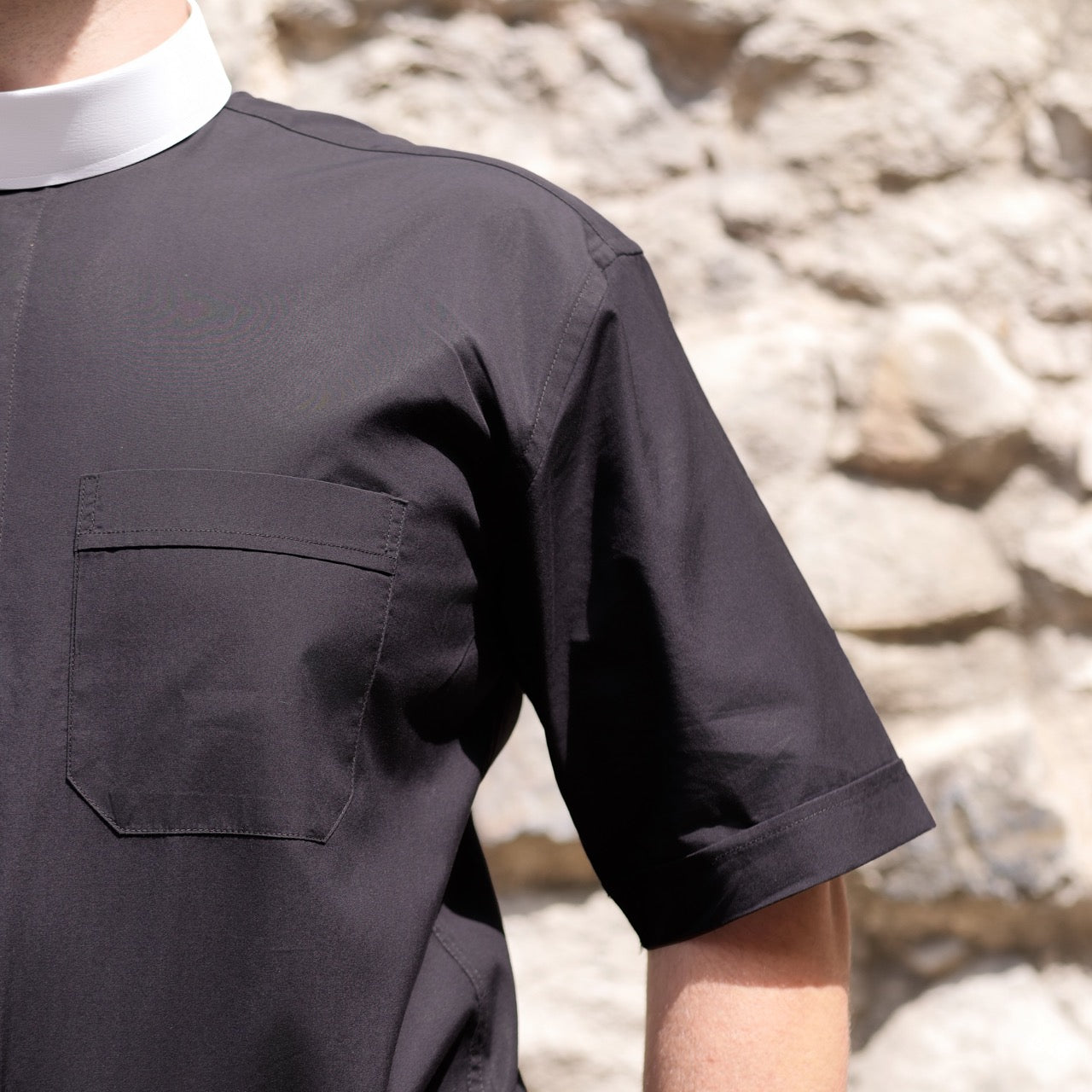 Close-up of a black tunic-style clergy shirt with a white collar, showcasing the pocket and fabric details.